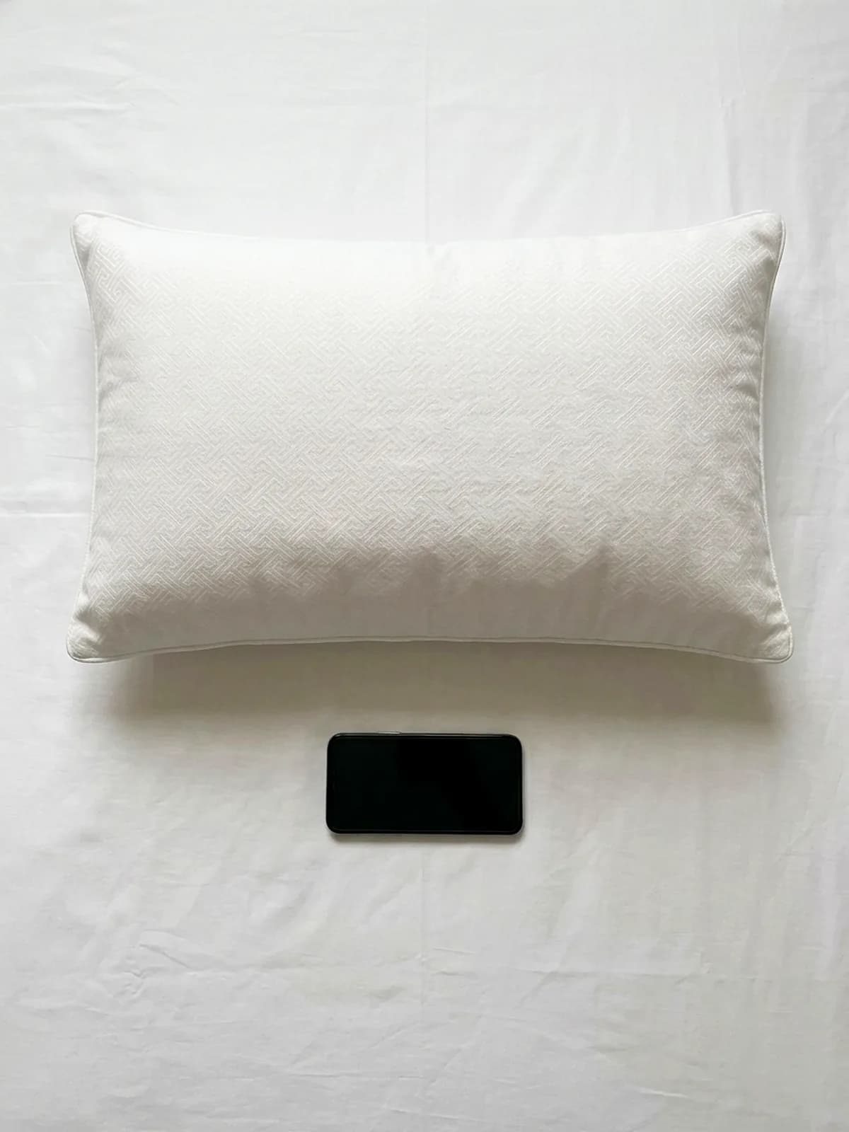 Una almohada de 40 por 60 centímetros vista desde arriba sobre una cama tendida, con un teléfono celular al lado para comparar el tamaño.